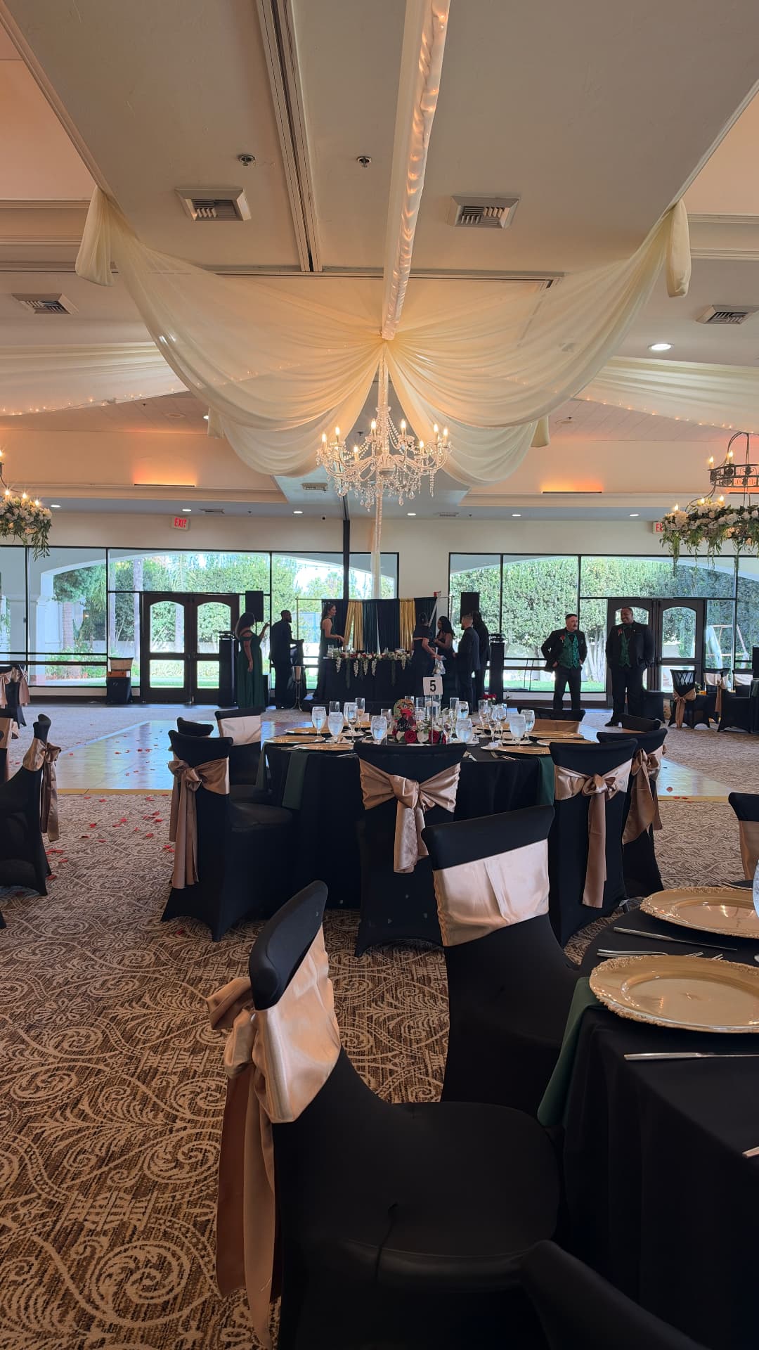A wedding reception ballroom with ivory ceiling drapery, a crystal chandelier, and tables set in black and gold