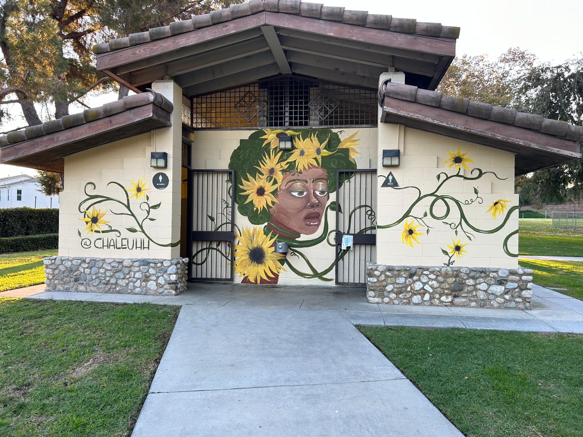 A park building painted with a woman crowned in sunflowers and green vines, signed @CHALEUHH