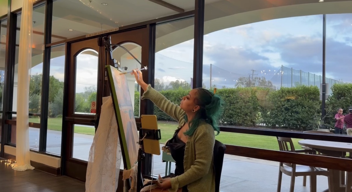 Chaleuh painting at her easel beside tall windows at a wedding venue