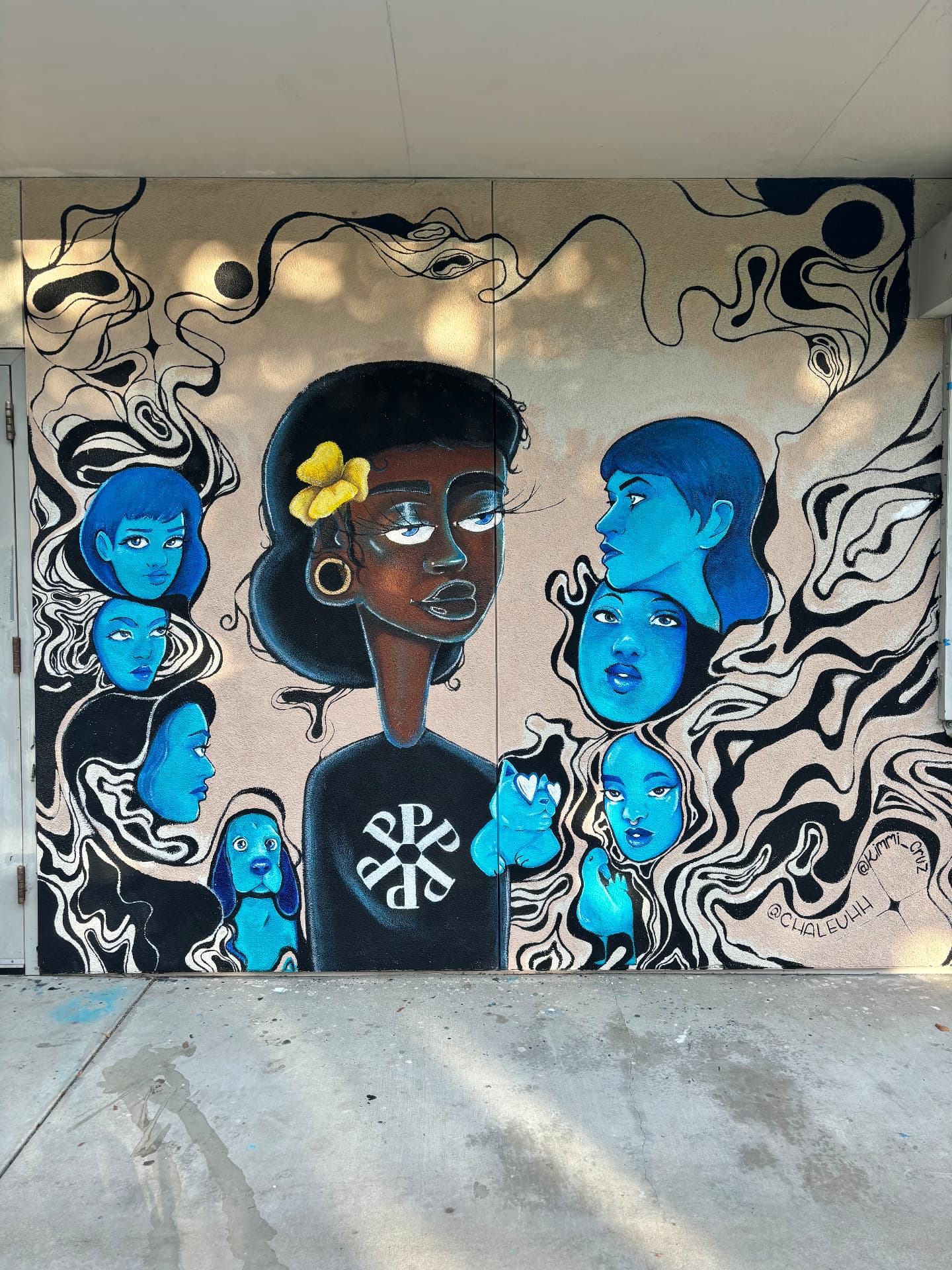 A mural of a central portrait with a yellow flower, surrounded by blue faces and flowing black and cream lines