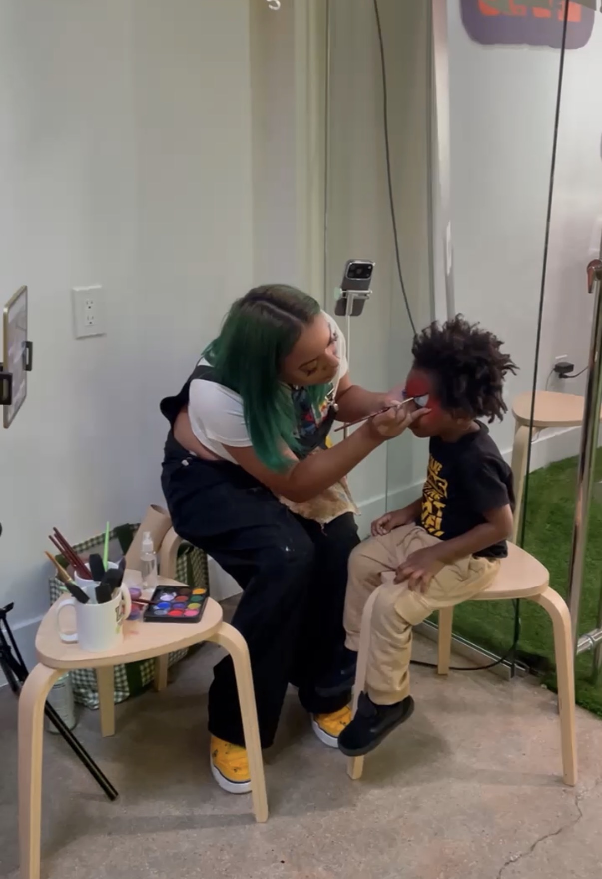 Chaleuh at her face painting station with a young guest on a stool and her paint palette beside them