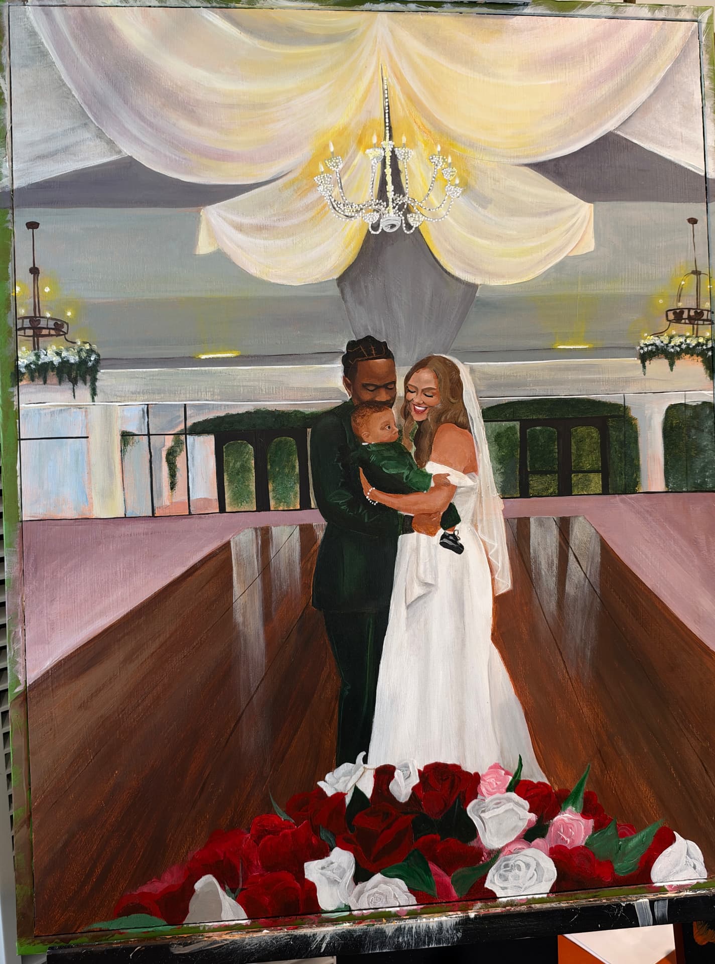 A live wedding painting of a bride and groom holding their baby beneath a draped chandelier, with roses in the foreground