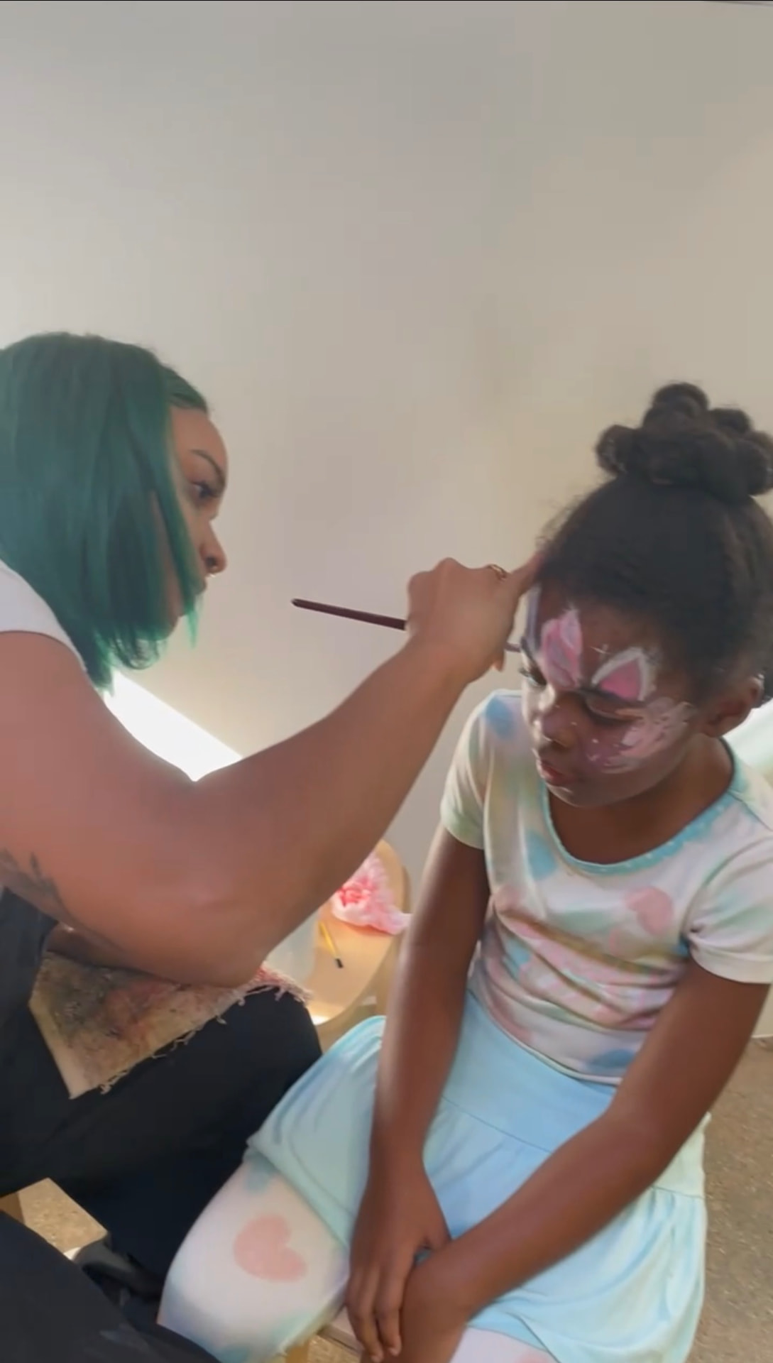 A young guest sitting still as Chaleuh adds detail to a pink and white butterfly face design